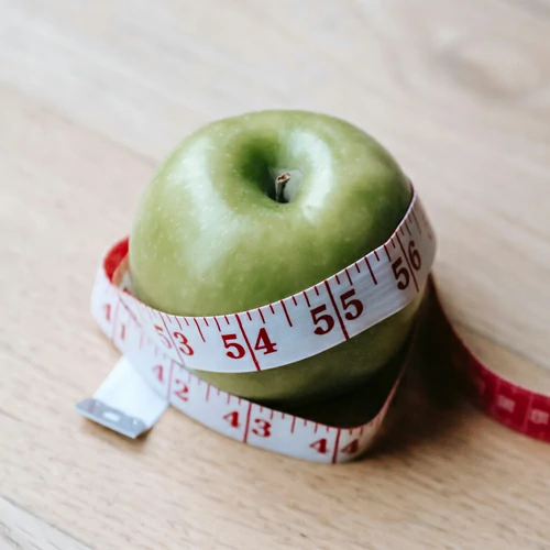 Una imagen en la que se ve una manzana verde enrollada con una cinta métrica amarilla, sobre una mesa de madera clara, representando la idea de comer sano y cuidar la peso en el trabajo.
