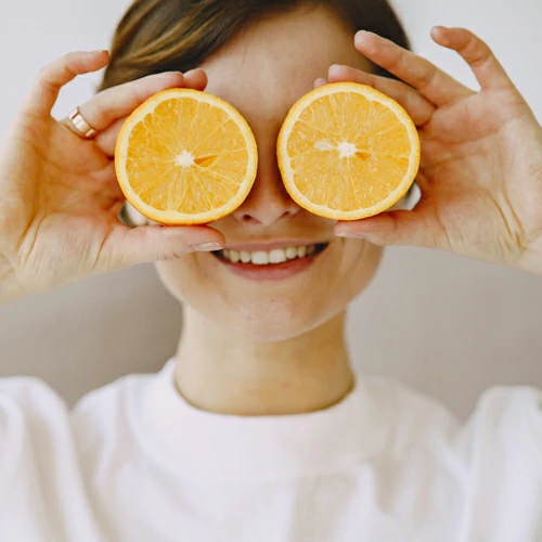 Una mujer joven y alegre, con el pelo rubio recogido en una coleta, sostiene dos rodajas de naranja fresca tapando sus propios ojos, sobre un fondo blanco.