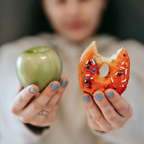 Una imagen en la que se ve una manzana y un donut glaseado en las manos estiradas de una mujer difuminada al fondo, representando la elección entre una opción saludable y una opción tentadora para el desayuno en la oficina.
